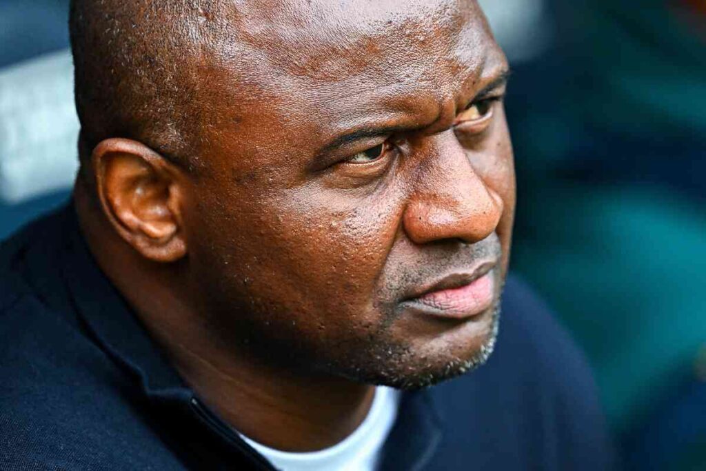 Patrick Vieira osserva concentrato dalla panchina durante una partita della stagione 2025, espressione intensa e sguardo fisso sul campo.